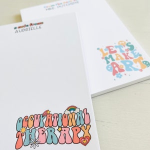 Peut inclure: Deux blocs-notes blancs avec des motifs color&eacute;s. Un bloc-notes a le texte "Let's Make Art" dans une police arc-en-ciel avec des fleurs et des visages souriants. L'autre bloc-notes a le texte "Occupational Therapy" dans une police arc-en-ciel avec un arc-en-ciel, une &eacute;toile, une fleur et un c&oelig;ur.