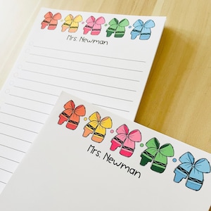 May include: Two white notepads with lined paper. The top of each notepad has a row of colorful crayons tied with bows. The text "Mrs. Newman" is printed on each notepad.