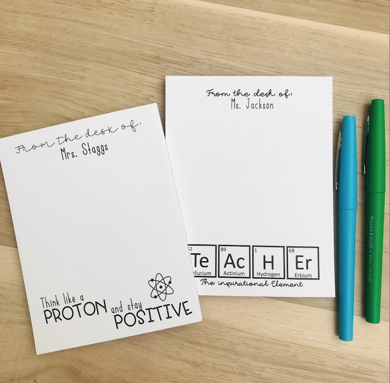 以下が含まれることがあります： 「From the desk of: Mrs. Staggs」と「From the desk of: Ms. Jackson」という黒い文字が書かれた白いメモ帳が2つあります。左側のメモ帳には、原子を黒と白で描いたイラストと「Think like a PROTON and stay POSITIVE」という文字があります。右側のメモ帳には、Te、Ac、H、Erの元素が強調表示された黒と白の周期表があります。周期表の下のテキストには「The inspirational element」と書いてあります。