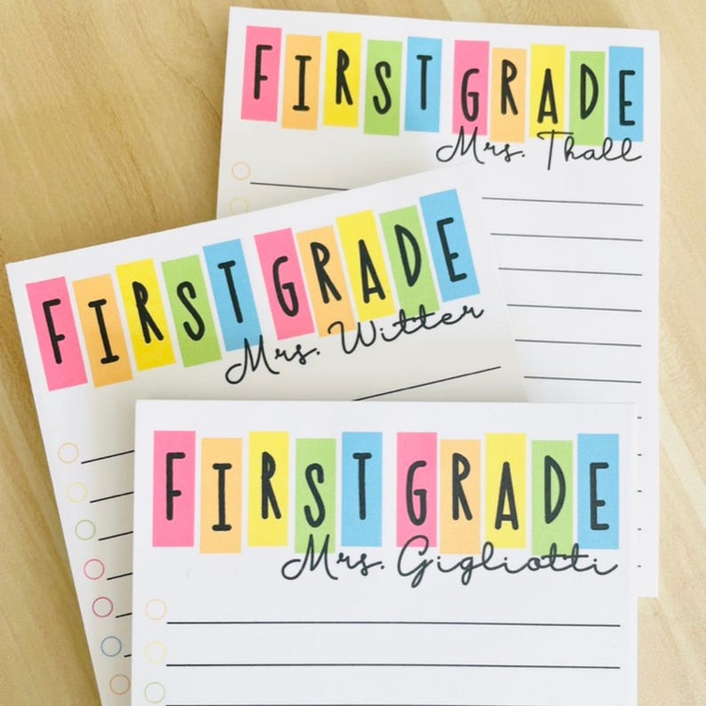 May include: Three white notepads with colorful stripes and the text "FIRST GRADE" in black. Each notepad has a different teacher's name: "Mrs. Thall", "Mrs. Witten", and "Mrs. Gigliotti".