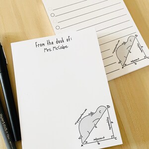 May include: A white notepad with the text From the desk of: Mrs. McCabe and a cartoon illustration of a hippopotamus forming a right triangle with labels for hypotenuse, opposite, and adjacent sides. A black pen is positioned to the left.