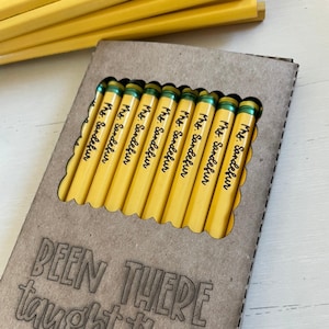 May include: A box of yellow pencils with green accents, each pencil is labeled "Mrs. Sandefur". The box is made of brown cardboard and has the text "BEEN THERE taught that" printed on the front.