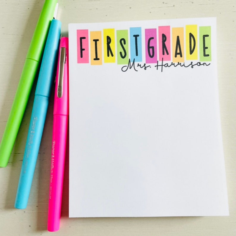 May include: A white notepad with the words "FIRST GRADE" in colorful block letters and the text "Mrs. Harrison" in black cursive writing. The notepad is sitting on a white surface with three highlighters.