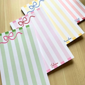 May include: Three rectangular stationery notepads featuring vertical stripes in pastel green, pink, and yellow. Each notepad displays a hand-drawn bow graphic at the top and a personalized name printed in cursive script at the bottom, reading Jolene, Annalee, and Juliet.