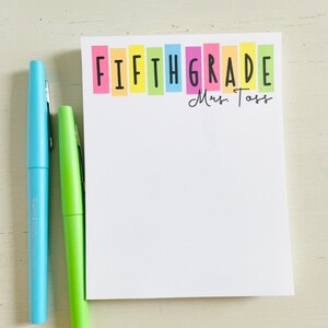 May include: A white notepad with the words "FIFTH GRADE" in black letters on a rainbow background. The name "Mrs. Toss" is written in black cursive below the rainbow.