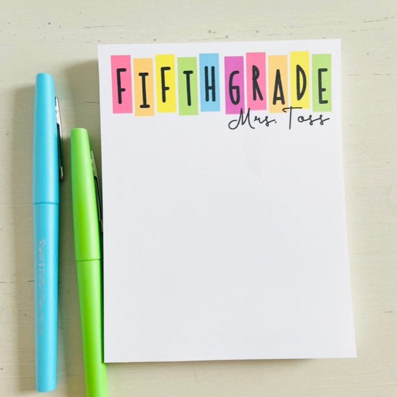 May include: A white notepad with the words "FIFTH GRADE" in black letters on a rainbow background. The name "Mrs. Toss" is written in black cursive below the rainbow.
