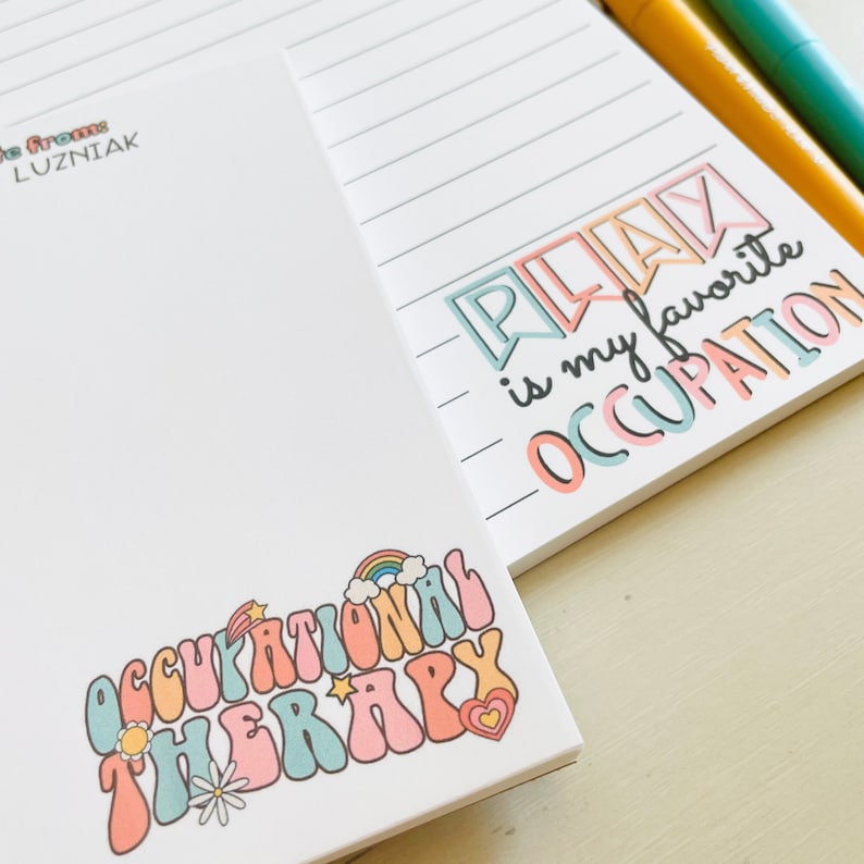 Peut inclure: Un bloc-notes avec une page lign&eacute;e et un graphique color&eacute; qui dit "Jouer est mon occupation pr&eacute;f&eacute;r&eacute;e". L'autre c&ocirc;t&eacute; du bloc-notes a un graphique color&eacute; qui dit "Th&eacute;rapie occupationnelle" avec un arc-en-ciel et des &eacute;toiles.