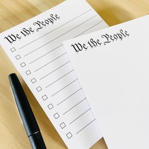 May include: Two white notepads with black lines and the text "We the People" in a decorative font. A black pen is lying on the top notepad.