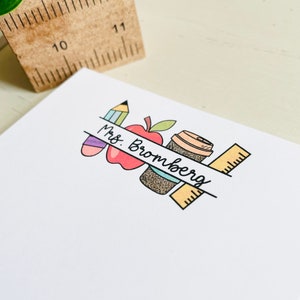 May include: A white notepad with a colorful illustration of a pencil, apple, coffee cup, and ruler. The text "Mrs. Bromberg" is written on a banner across the illustration.
