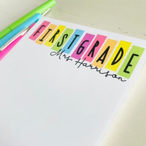 May include: A white notepad with the words "FIRST GRADE" in black letters on colorful rectangular strips. The text "Mrs. Harrison" is written in black cursive below the word "GRADE".