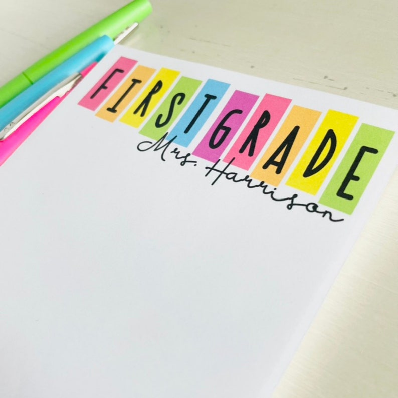 May include: A white notepad with the words "FIRST GRADE" in black letters on colorful rectangular strips. The text "Mrs. Harrison" is written in black cursive below the word "GRADE".