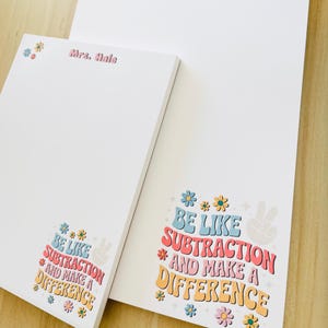 Könnte beinhalten: Zwei weiße Notizblöcke mit farbenfrohem Text und floralen Designs. Auf einem Notizblock steht "Mrs. Hale" in Rosa. Der andere Notizblock lautet "BE LIKE SUBTRACTION AND MAKE A DIFFERENCE" in Retro-Farben. Die Notizblöcke haben eine Vintage-Ästhetik.
