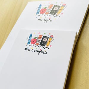May include: Two white rectangular notepads featuring a colorful illustration of a blue crayon, a red apple, an orange glue stick, a black notebook, and a yellow pencil with faces and limbs. The top pad says Ms. Apple and the bottom says Mrs. Campbell.