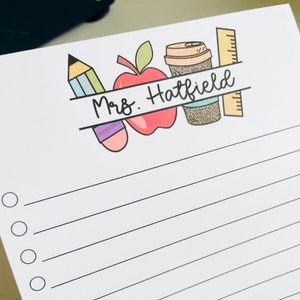 May include: A white notepad with a black and white line design. The top of the notepad has a banner with the text "Mrs. Hatfield" and illustrations of a pencil, apple, and coffee cup.
