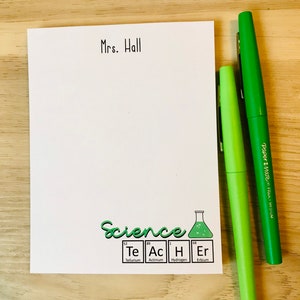以下が含まれることがあります： 「Mrs. Hall」というテキストが上部に書かれた白いメモ帳。メモ帳の下部には、「Science」という単語と、テルル、アクチニウム、水素、エルビウムの化学記号が書かれた緑色の科学をテーマにしたデザインが施されています。