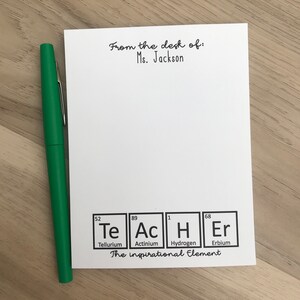 以下が含まれることがあります： 「From the desk of: Ms. Jackson」という黒い文字が書かれた白いメモ帳。テキストの下には、Te、Ac、H、Erの4つの化学元素の図があります。元素の下のテキストは「The inspirational Element.」です。
