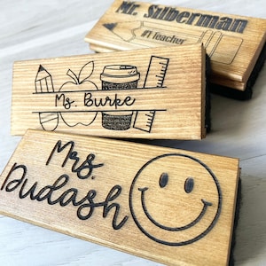 May include: Three wooden chalkboard erasers with personalized teacher names. The top eraser says "Mr. Silberman #1 Teacher". The middle eraser says "Ms. Burke" with a pencil, apple, and coffee cup design. The bottom eraser says "Mrs. Dudash" with a smiley face.