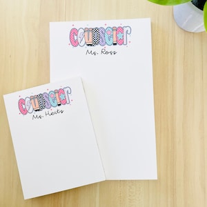 May include: Two white notepads with the word "Counselor" in colorful bubble letters. The larger notepad has "Ms. Ross" printed below the word, and the smaller notepad has "Ms. Heitz" printed below the word.