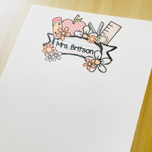Może przedstawiać: Biały papier listowy z kwiatowym wzorem i napisem "Mrs. Britson". Projekt zawiera ołówek, jabłko, nożyczki, linijkę i kwiaty w odcieniach różu, brzoskwini i bieli. Papier listowy jest pusty, z białym tłem.