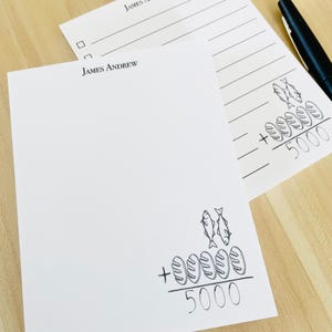 May include: A black and white drawing of two fish and six loaves of bread with the number 5000 written below. The drawing is on a white notepad with the words "JAMES ANDREW" printed at the top.