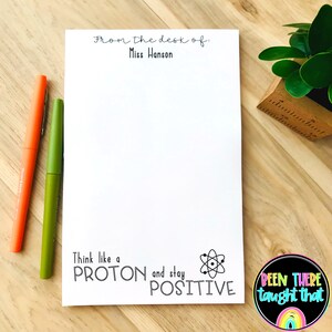以下が含まれることがあります： 「From the desk of: Miss Hanson」と黒文字で書かれた白いメモ帳。メモ帳には、原子を黒と白で描いたイラストと、「Think like a PROTON and stay POSITIVE」という文字が書かれている。メモ帳は木製の表面に置かれ、2本のペンが置かれている。
