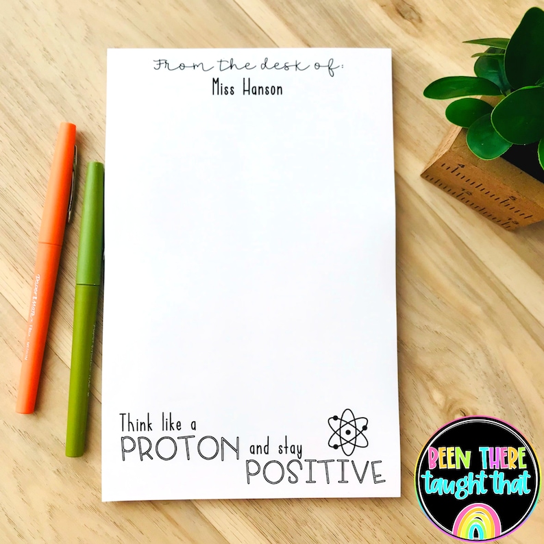 以下が含まれることがあります： 「From the desk of: Miss Hanson」と黒文字で書かれた白いメモ帳。メモ帳には、原子を黒と白で描いたイラストと、「Think like a PROTON and stay POSITIVE」という文字が書かれている。メモ帳は木製の表面に置かれ、2本のペンが置かれている。