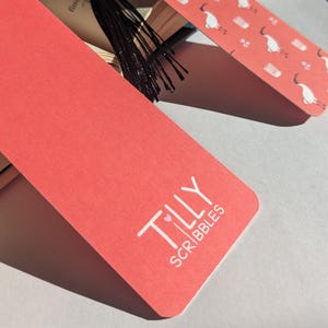 Pu&ograve; includere: Un segnalibro rosso con il testo "TILLY SCRIBBLES" in bianco con un cuore rosa tra le parole "TILLY" e "SCRIBBLES".