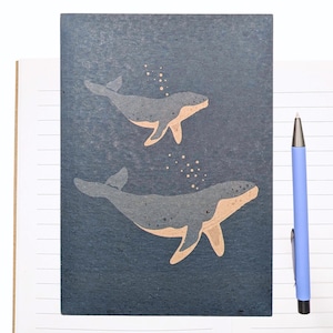 May include: A dark blue rectangular notebook cover featuring two illustrated whales in shades of grey and tan, with small bubbles rising above them. A light blue ballpoint pen with a silver tip and clip is positioned vertically to the right side.