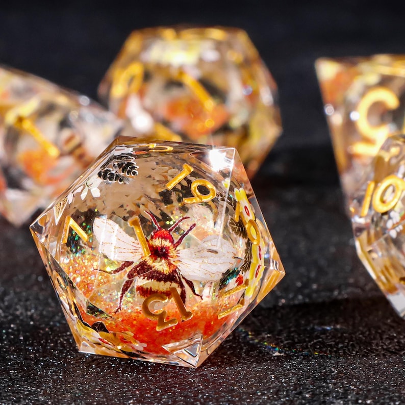 May include: A clear, multi-sided dice with a bee and floral design. The dice is filled with orange glitter and has gold numbers.