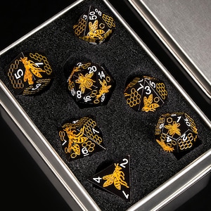May include: A set of seven black polyhedral dice with gold-coloured bee and honeycomb engravings, featuring white numbers. The dice are arranged in a silver-toned metal storage tin with a black foam insert that has custom-cut slots for each piece.