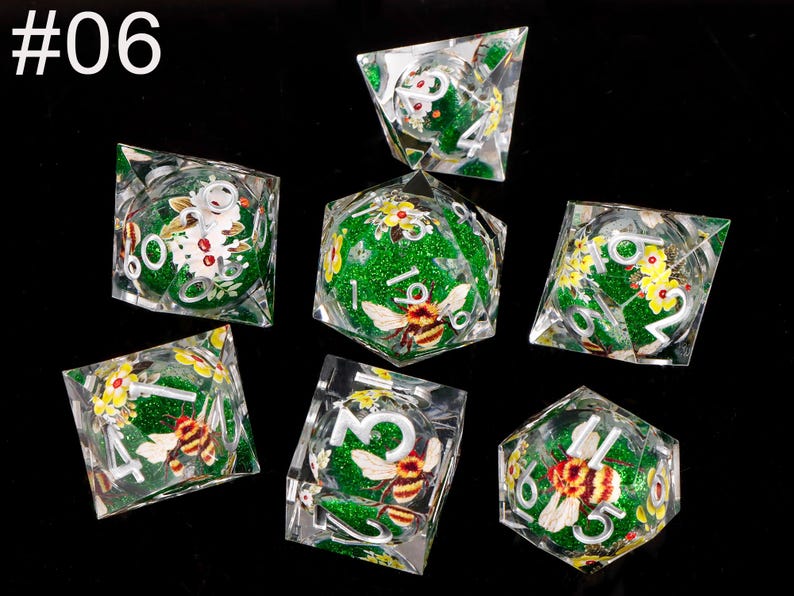 May include: Set of seven green and clear dice with a glitter finish. Each die features a bee and floral design. The dice are numbered 1 through 6.