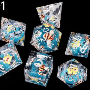 May include: Set of seven clear acrylic dice with blue and turquoise glitter, featuring a bee and floral design. The dice are numbered 1 through 6.