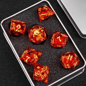 Puede incluir: Un conjunto de siete dados rojos y dorados con motas doradas y números. Los dados están en una caja plateada con acolchado de espuma negra.