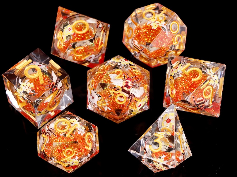 May include: Set of seven clear dice with orange glitter and gold accents. Each die has a different number from 1 to 7. The dice are decorated with white flowers and a bee.