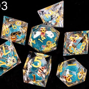 May include: Set of seven turquoise and gold dice with bee and flower designs. The dice are made of clear resin with a glittery finish. The numbers on the dice are in gold.