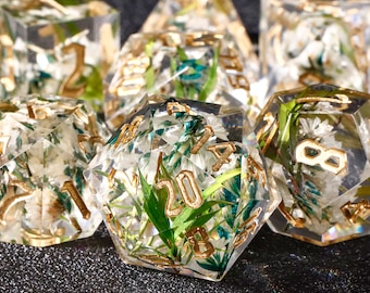 May include: A set of seven clear resin dice with gold numbers and embedded dried flowers and greenery. The dice are in various shapes, including a d20, d12, d10, d8, d6, and d4.