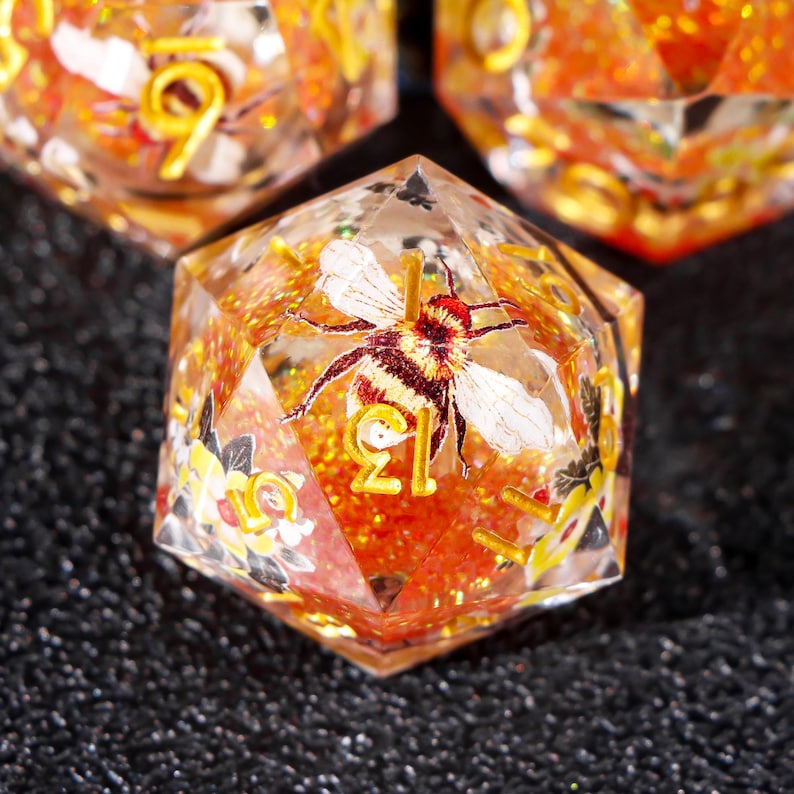 May include: A clear, orange, and gold-accented dice with a bee illustration and glitter. The dice is a 12-sided die with the number 12 visible.