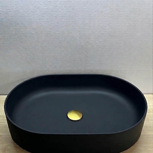 Peut inclure: Un lavabo de salle de bain noir mat, de forme ovale, avec un couvercle de drain doré. Le lavabo est posé sur une surface en bois clair, avec un mur gris clair en arrière-plan. Le lavabo a un design moderne et minimaliste.