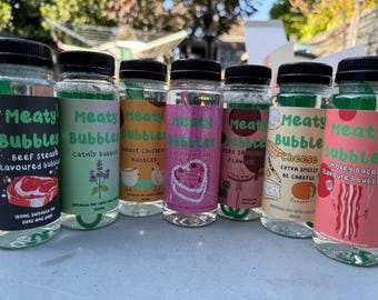 May include: Eight clear plastic bottles with green lids and labels that say "Meaty Bubbles" in green text. Each bottle has a different flavour: Beef Steak, Catnip, Roast Chicken, Birthday Cake, Pork Sausage, Cheese, and Smokey Bacon. The bottles are arranged in a row on a light brown surface.