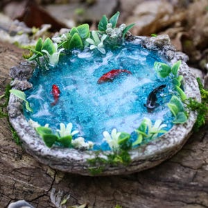 May include: A miniature pond scene with a blue water effect, featuring three koi fish in red and black. The pond is surrounded by green plants, white flowers, and a stone-like border. The scene is set on a wooden surface.