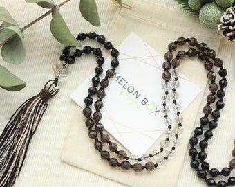 May include: A layered necklace with faceted black, clear, and smoky quartz beads. A brown and white tassel hangs from the necklace. A small card with the text "MELON BOX Designs" is visible.