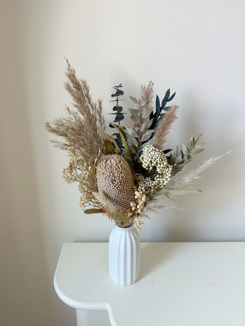 Puede incluir: Un jarr&oacute;n de cer&aacute;mica blanco y acanalado contiene un ramo de flores secas. El arreglo incluye hierba de la pampa beige, eucalipto y una gran protea beige texturizada. Otras flores secas y vegetaci&oacute;n a&ntilde;aden textura y color.