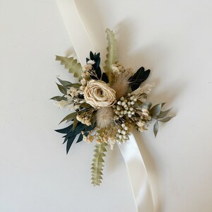 Puede incluir: Un ramillete de mu&ntilde;eca con una rosa color crema y varias flores secas en tonos verdes, beige y marr&oacute;n. El arreglo floral est&aacute; sujeto a una cinta de sat&eacute;n color crema. Perfecto para bodas o eventos especiales.