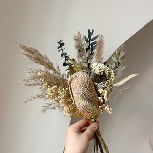 Puede incluir: Un ramo de flores secas con una gran flor de banksia beige texturizada como punto focal. El arreglo incluye hierba de la pampa, eucalipto y otros elementos secos en tonos neutros, atados con hilo.