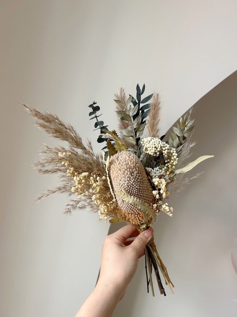 Puede incluir: Un ramo de flores secas con una gran flor de banksia beige texturizada como punto focal. El arreglo incluye hierba de la pampa, eucalipto y otros elementos secos en tonos neutros, atados con hilo.