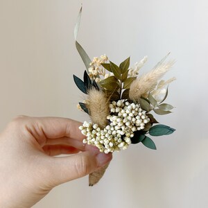 Puede incluir: Un peque&ntilde;o prendedor artesanal con una variedad de flores y vegetaci&oacute;n secas. El arreglo incluye elementos blancos, beige y verdes, envuelto con hilo de c&aacute;&ntilde;amo. El prendedor se sostiene en una mano sobre un fondo neutro.