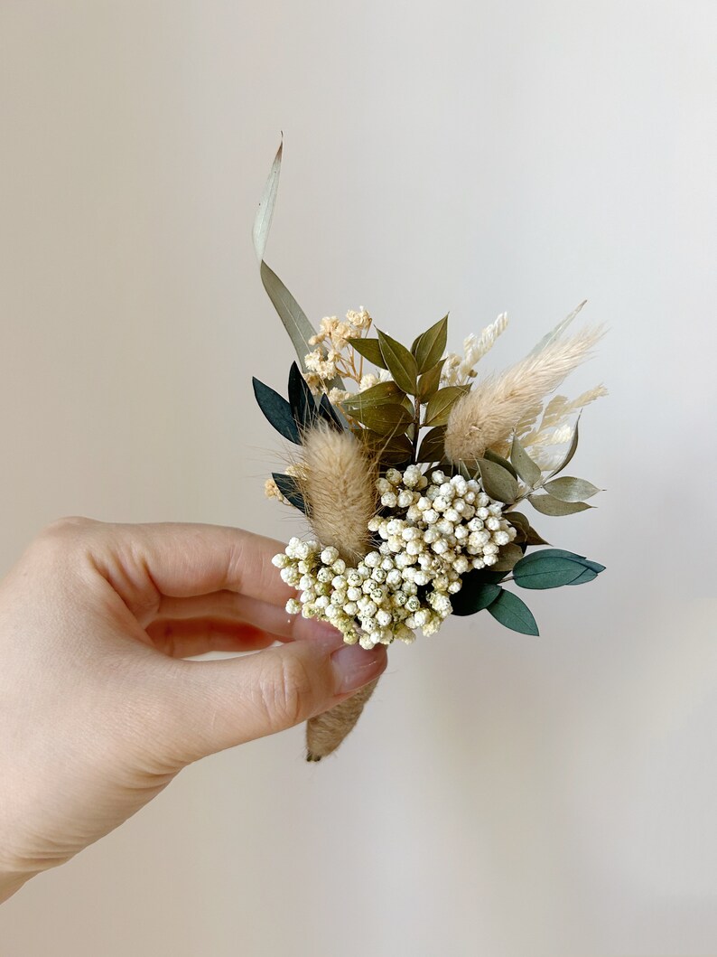 Puede incluir: Un peque&ntilde;o prendedor artesanal con una variedad de flores y vegetaci&oacute;n secas. El arreglo incluye elementos blancos, beige y verdes, envuelto con hilo de c&aacute;&ntilde;amo. El prendedor se sostiene en una mano sobre un fondo neutro.