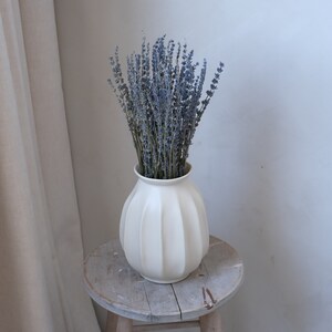 May include: A white, ribbed ceramic vase filled with dried lavender. The vase sits on a small, weathered wooden stool. The lavender has a muted purple hue, contrasting with the vase's creamy white color. The scene evokes a sense of rustic charm and natural beauty.