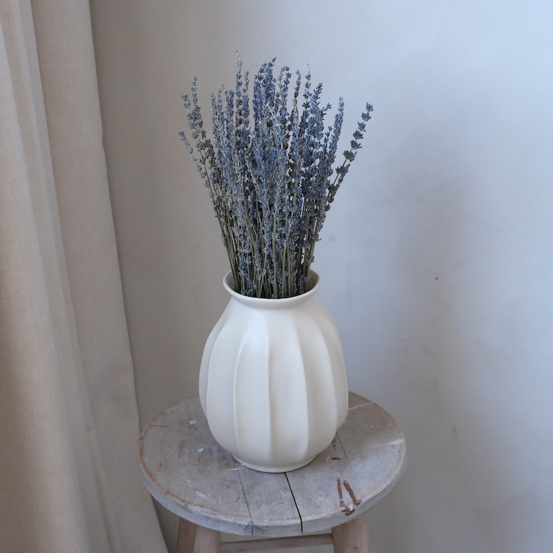 May include: A white, ribbed ceramic vase filled with dried lavender. The vase sits on a small, weathered wooden stool. The lavender has a muted purple hue, contrasting with the vase's creamy white color. The scene evokes a sense of rustic charm and natural beauty.