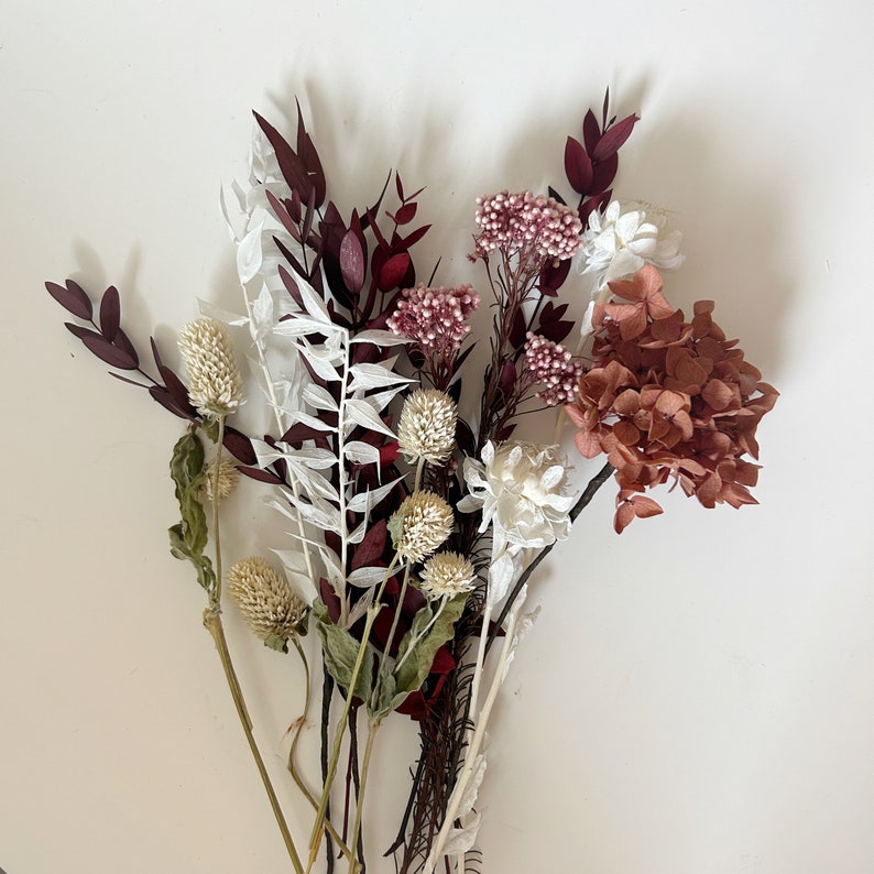 May include: A bouquet of dried flowers in shades of white, brown, and burgundy. The bouquet includes white and brown dried flowers, burgundy dried leaves, and a cluster of small, dried burgundy flowers.